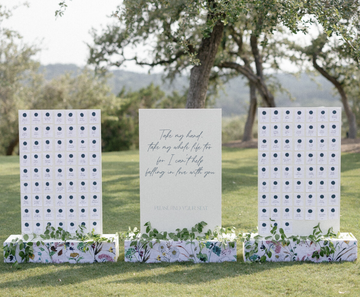 Modern outdoor wedding signage display with custom seating chart walls and romantic quote sign, accented with floral boxes.